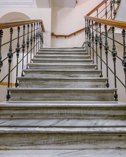 A straight marble staircase inside a building with a wooden handrail on each side and decorative black metal balustrades, leading upward and out of view. The stairs are illuminated by ambient light, h