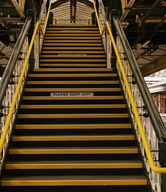 A straight marble staircase inside a building with a wooden handrail on each side and decorative black metal balustrades, leading upward and out of view. The stairs are illuminated by ambient light, h