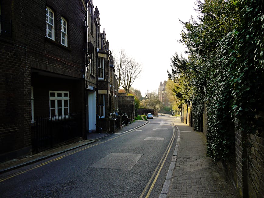 A quiet residential street in Hampstead, showing a narrow pavement beside a row of dark brick terraced houses with white-framed windows and small front gardens enclosed by black wrought iron fences. The street is partially shaded by leafless and green trees, with a parked white van visible in the distance, suggesting ongoing household furniture transportation or home relocation activities. The sidewalk is lined with paving stones, and the road has yellow double lines along the curb, indicating parking restrictions. This scene is typical of an area serviced by local house removals and moving companies such as Man and Van Hampstead, which offers professional relocation and furniture transport services, demonstrating a typical location for loading or unloading items during a house move in a tranquil neighbourhood setting.