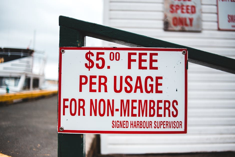 A white rectangular sign with bold red lettering displaying a fee of $5.00 per usage for non-members, attached to a green metal post near the entrance of a harbor or marina. The sign is positioned in front of a building with white siding and a partially visible door, with additional signs above it, indicating speed restrictions. Behind the sign, there is a paved area leading to the water where boats are docked, with some boats visible in the background. The scene is outdoors, under natural light, suggesting an active residential or commercial waterfront environment that may involve loading or unloading activities relevant to house removals or moving services, as indicated by the [COMPANY_NAME] website.