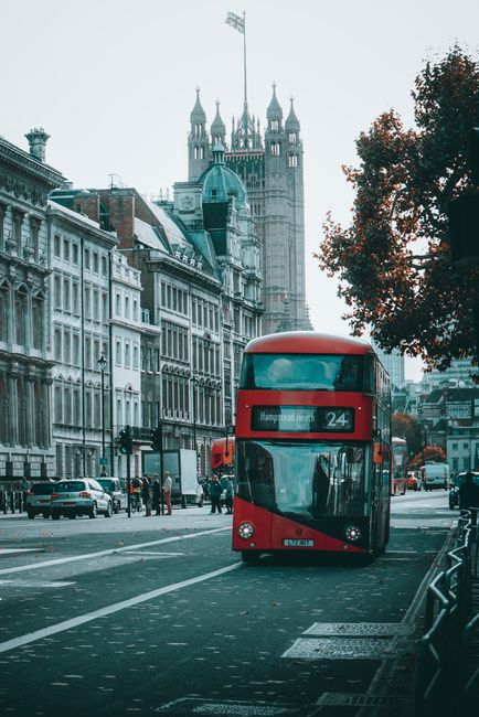 A red double-decker bus displaying the route number 24 and destination Hampstead on the front, is stationary on a busy street in London with pedestrians walking along the pavement and cars parked beside it. Behind the bus, there is a row of traditional London-style buildings with detailed facades and large windows, and in the background, the iconic tower of Westminster Palace with its spires and clock tower is visible under overcast skies. The scene is typical of central London and captures the urban environment where home relocation and furniture transport services by Man and Van Hampstead may be utilized during house moves, with the bus suggesting a connection to the area's public transport network as part of moving logistics.