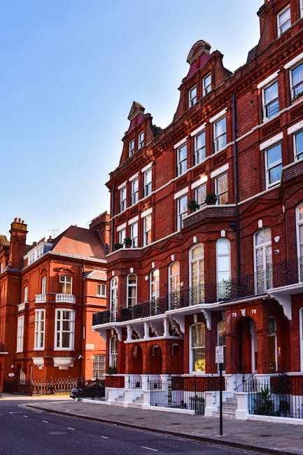 Image of a multi-storey red brick residential building on Heath Street Hampstead, showing several large windows with some having black wrought iron balconies, and white decorative elements around the windows and at the building's base. The building features a curved corner with a prominent rounded roof detail at the top. It is situated on a quiet urban street with a paved pavement and a few parked cars visible. The sky above is clear and blue, indicating a bright day. The scene exemplifies typical architecture in Hampstead, suitable for home relocation or furniture transport services, as provided by Man and Van Hampstead. The image captures the exterior environment where moving and packing activities might take place during house removals, emphasizing the accessibility of the property for loading and unloading furniture and boxes.
