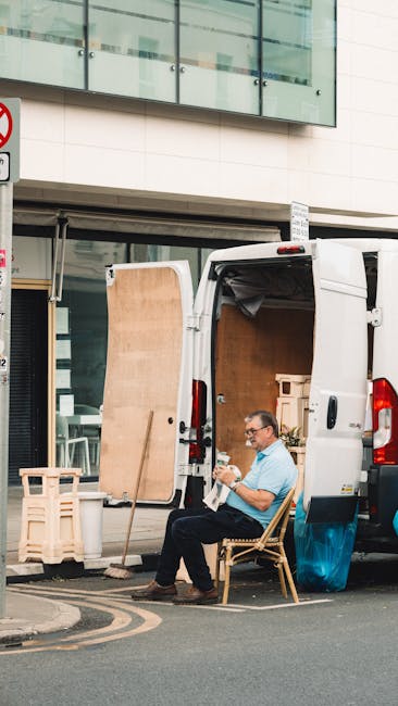 A man sitting on a wooden chair next to the open rear doors of a white moving van parked on a city street in Hampstead, South End Green, engaged in writing or reading. Inside the van, visible items include large cardboard boxes, wooden panels, and wrapped furniture components prepared for a house removal or small business move. The van is positioned partially on the pavement with the open loading area revealing packing materials and shipment essentials, while a building with glass windows and architectural features is seen above. The scene depicts a packing and loading process as part of a professional relocation service, with additional packing supplies such as protective blankets and wrapping materials nearby, consistent with furniture transport and move logistics associated with [PAGE_TITLE]. Man and Van Hampstead provides removals services, as indicated by the professional setup and arrangement of items for transport.