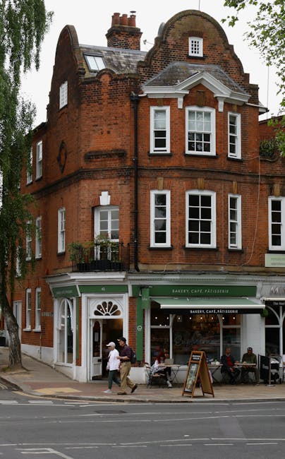 The image depicts a three-story red brick building on a street corner, with the ground floor housing a bakery, cafe, and pâtisserie, indicated by a green awning with white lettering. Large display windows showcase the shop interior, and a wooden sandwich board signs a promotion on the sidewalk. Outside the bakery, there are four people sitting at outdoor tables, with one person standing nearby. Two pedestrians, one wearing a white shirt and the other in a dark jacket, are walking past the shop. The upper floors feature several white-framed sash windows, some with small balconies and ornamental brickwork, and the building has a slightly curved gabled roof with chimney stacks. The surroundings include a tree with lush green foliage partially visible on the left side, and the scene is illuminated by natural daylight, suggesting a bright, clear day. As part of a house or business relocation process, Man and Van Hampstead might use such a setting to load furniture or boxes during a local move, particularly in Hampstead’s South End Green area, within their removals services.