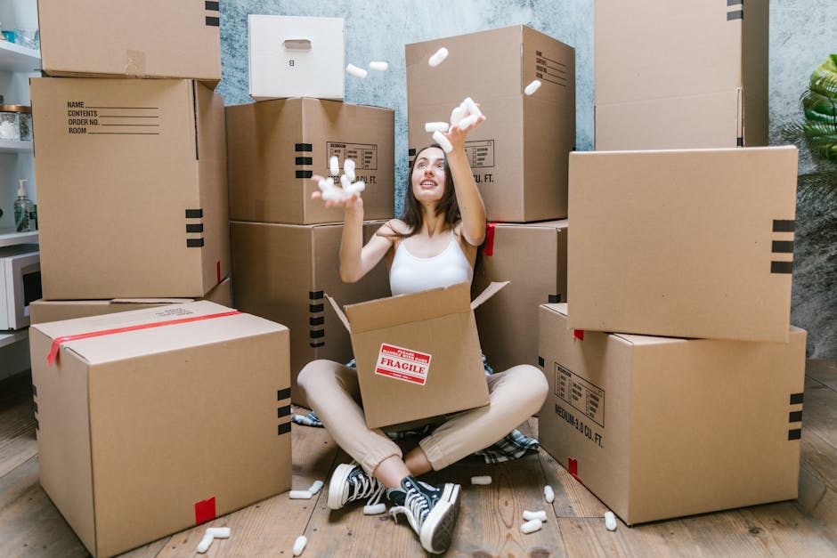 A young woman with long dark hair, sitting cross-legged on a wooden floor inside a room, surrounded by multiple cardboard moving boxes of various sizes, some sealed with red packing tape and marked with fragile labels. She is wearing a white tank top, beige pants, and black sneakers with white soles. She is smiling and tossing white packing peanuts into the air, with some peanuts falling back into a partially open box in her lap. The background includes a textured blue wall, a green potted plant on the right, and an area of a countertop on the left with a small appliance, indicating she is in a home environment during a home relocation process managed by Man and Van Hampstead, as seen on the website page for packing and removals Hampstead Heath specialist tips.