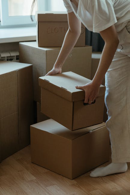 A person stacking and organizing brown cardboard boxes in an indoor space, likely preparing for a house removal or relocation. The boxes vary in size and are labeled, with one marked 'CLOTHES,' indicating packing of personal belongings. The individual is partially visible, wearing light-colored clothing and white socks, using both hands to handle a box positioned on top of others. The environment features natural light coming through a window, with a wooden floor and additional boxes arranged nearby, suggesting an ongoing packing or moving process. This scene is associated with home relocation services provided by Man and Van Hampstead, including packing and furniture transport, and takes place within a domestic setting as part of a professional removals operation.