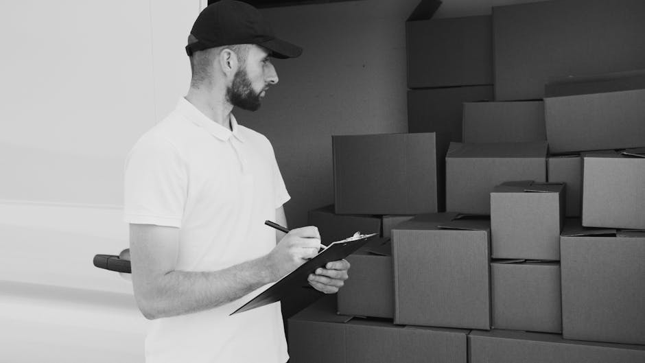 A man wearing a white polo shirt and a black cap stands indoors beside a stack of uniformly sized cardboard boxes, some of which are taped closed. He holds a clipboard and a pen, appearing to take not