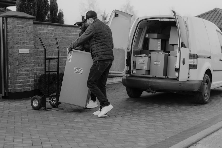 A man wearing a white polo shirt and a black cap stands indoors beside a stack of uniformly sized cardboard boxes, some of which are taped closed. He holds a clipboard and a pen, appearing to take not